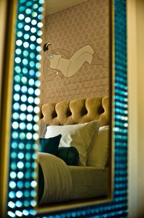 Bed with teal cushions and an Art Deco wall drawing reflected in a turquoise crystal-framed mirror