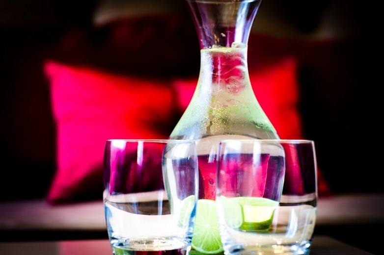 Carafe of water with lime and two tumblers on a table, ruby-red cushions behind